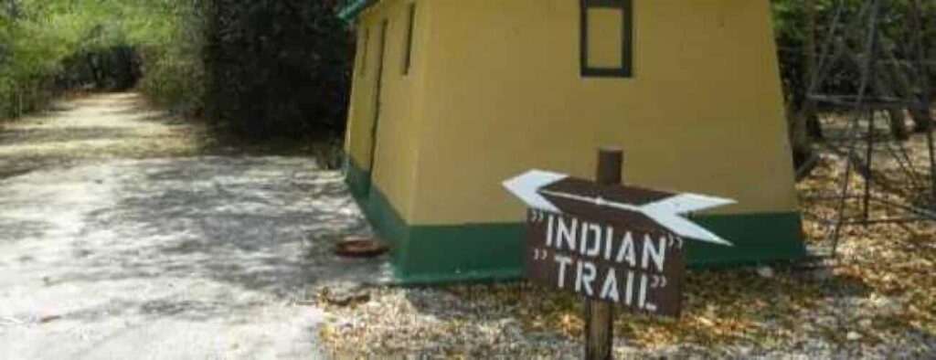 A dirt path runs past a small yellow and green building. In front, a wooden sign with white letters and arrows points right, marking the Indian Trail. Trees line the path, with dry leaves scattered on the ground. Product name: Indian Trail.