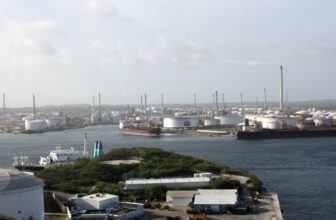 A large oil refinery complex is seen by the Curacao waterfront, with storage tanks, tall chimneys, and smokestacks. Several ships and tankers are docked nearby, while vegetation and smaller buildings appear in the foreground under a mostly cloudy sky.