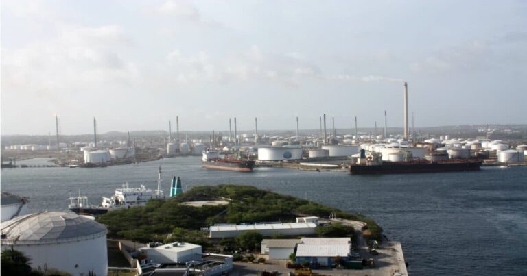 A large oil refinery complex is seen by the Curacao waterfront, with storage tanks, tall chimneys, and smokestacks. Several ships and tankers are docked nearby, while vegetation and smaller buildings appear in the foreground under a mostly cloudy sky.