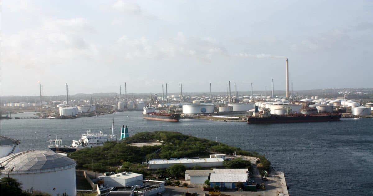 A large oil refinery complex is seen by the Curacao waterfront, with storage tanks, tall chimneys, and smokestacks. Several ships and tankers are docked nearby, while vegetation and smaller buildings appear in the foreground under a mostly cloudy sky.