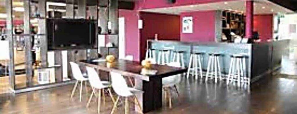 Een modern café-interieur met donkerhouten vloeren, een lange tafel en witte stoelen - perfect om te ontspannen na het gebruik van uw Langdurig sport lidmaatschap - plus een bar, rode accentmuren, een gemonteerde tv en glazen scheidingswanden met decoratieve voorwerpen. Goed verlicht en eigentijds.