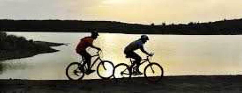 Experience mountain bike tours along a dirt path beside a calm lake at sunset, where two riders’ silhouettes stand out against glowing waters with rolling hills and a clear sky in the background.