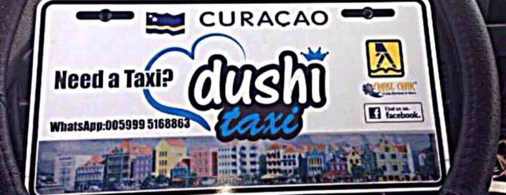 A Curaçao taxi sign for "Taxi to Playa Kenepa" features “Need a Taxi? dushi taxi” with a heart outline, WhatsApp +5999 516 6886, taxi and Facebook logos, and Willemstad art—ideal for rides to Playa Kenepa.