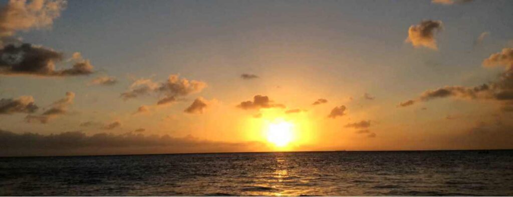 Experience the Relaxing Sun Set Trip: Watch a vibrant orange and yellow sun sink low over the ocean, as gentle waves ripple in the foreground and scattered clouds reflect warm light, creating a tranquil evening scene.