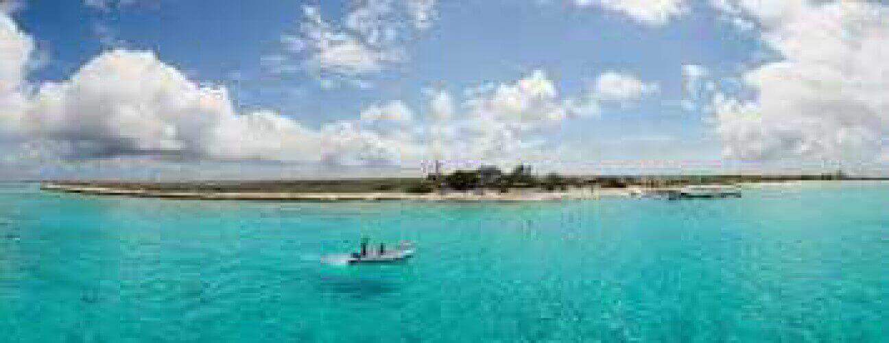 Two people in a small boat float on clear turquoise water near a sandy island with sparse plants, while another Rent a Yacht vessel is docked by the shore under a sky filled with fluffy clouds.