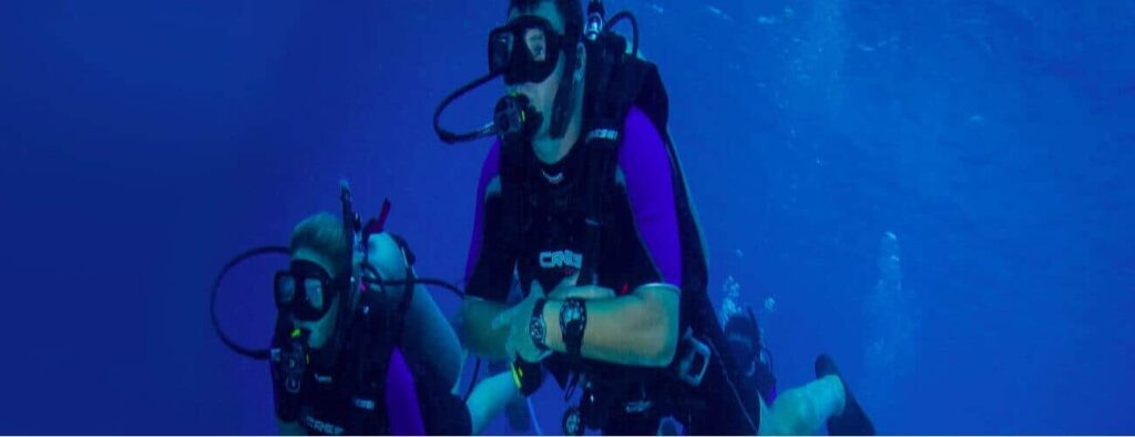 Two scuba divers in purple wetsuits explore the clear blue ocean, possibly practicing skills from the PADI Rescue Diving Course, surrounded by deep water and shimmering bubbles.