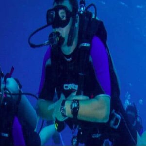 Twee duikers in zwarte wetsuits zwemmen onder water in de helderblauwe zee tijdens een PADI Cursus Reddingsduiken. Beiden dragen maskers en mondstukken terwijl bellen opstijgen en zonlicht door het water om hen heen glinstert.