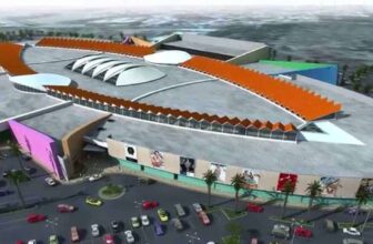 Aerial view of a large, modern Curacao shopping mall with a unique, oval-shaped design, colorful exterior walls, and orange zigzag roof accents. The parking lot below is filled with cars and bordered by palm trees; the surrounding area is blurred.