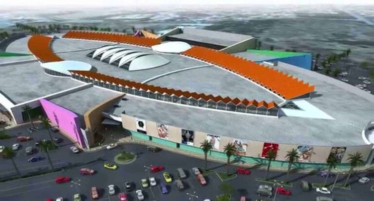 Aerial view of a large, modern Curacao shopping mall with a unique, oval-shaped design, colorful exterior walls, and orange zigzag roof accents. The parking lot below is filled with cars and bordered by palm trees; the surrounding area is blurred.