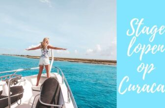A woman stands at the bow of a boat with arms outstretched, facing Curacao’s clear blue water and a distant shoreline under a partly cloudy sky. On the right, a light blue box displays white cursive text: Stage lopen op Curaçao.