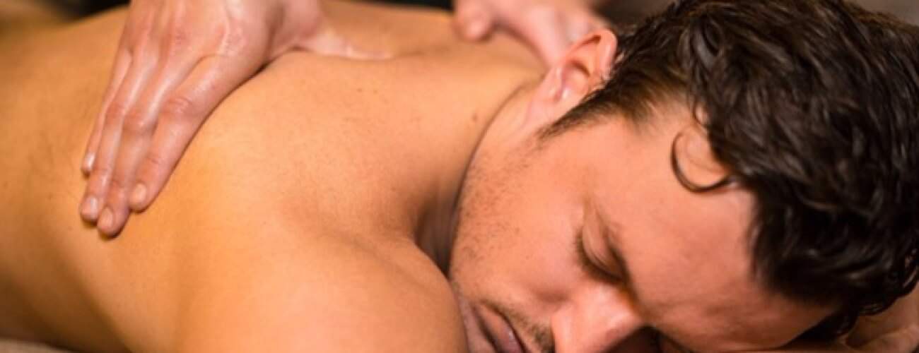 A man with dark hair lies face down with eyes closed, looking relaxed as he receives a Swedish Massage. The close-up highlights his upper body and the masseur’s hands skillfully massaging his back.