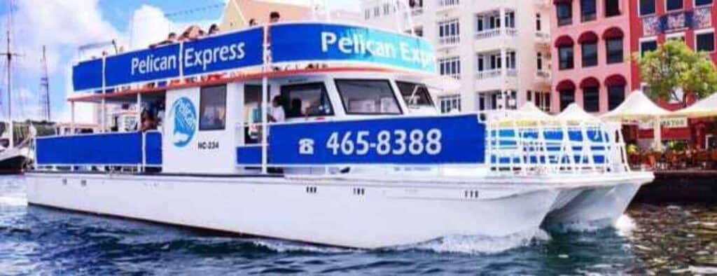 A white and blue catamaran named Pelican Express cruises past The Happy Harbour, a popular seafood spot by colorful waterfront buildings, displaying the phone number 465-8388 under a clear sky.