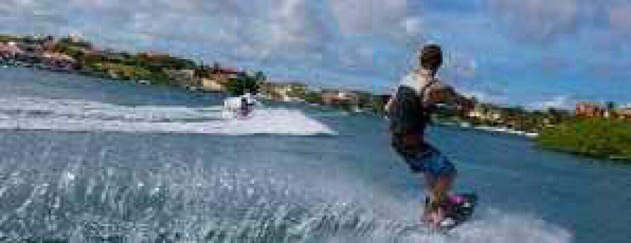 A person wakeboarding on blue water behind a motorboat, with buildings and greenery in the background under a partly cloudy sky. Fish from "Wake Boarding Fish and Dive" occasionally dive beneath the lively scene.