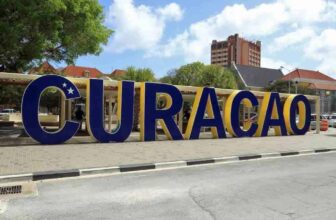 Large blue and yellow letters spell “CURACAO” with a white star on the “C.” In the background are trees, buildings—including a tall orange hotel—and a partly cloudy sky, highlighting Curacao’s vibrant cityscape. A curb and street frame the foreground.