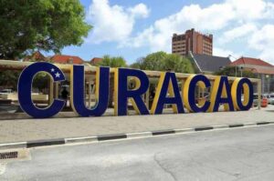 Large blue and yellow CURACAO letters stand outdoors, featuring a white star and crescent on the C. Set in a lively public square, this Curacao landmark is surrounded by trees, people, and tall buildings under a partly cloudy sky.