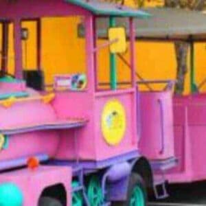 The Willemstad Tour by Trolley Train features a brightly colored pink and turquoise train with open passenger cars, parked on a paved road beside a yellow wall, creating a cheerful, playful vibe for your sightseeing adventure.