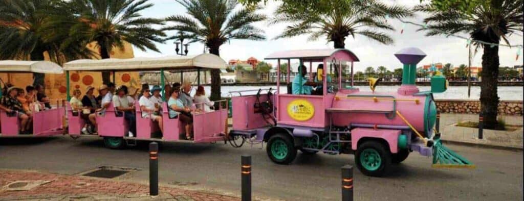 The Willemstad Trolley Train Tour features a vibrant purple and pink train providing a scenic ride along the palm-lined waterfront, where passengers in white-roofed cars enjoy city, water, and sky views.