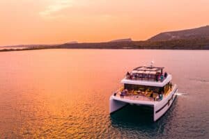 De Catamaran zeiltocht bij zonsondergang op Curaçao biedt een ontspannen tocht op een moderne witte catamaran met uitzicht op groene heuvels en de kustlijn, terwijl de lucht roze-oranje kleurt en passagiers genieten op het dek.