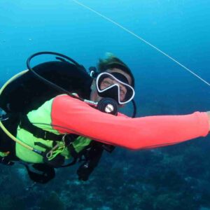 Wearing a neon green vest and red wetsuit, a scuba diver extends an arm underwater holding a device amid bubbles, with coral and blue ocean in the background—enjoy the 6-Day Unlimited Shore Diving Package.