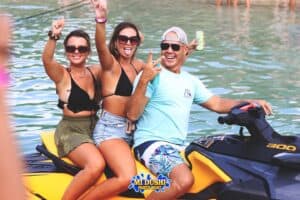 Three people smile and make peace signs while riding a yellow jet ski on the water. Two women in swimsuits and sunglasses sit behind a man in a blue shirt and cap. The Paranda Boat Trips in CuraƧao logo is at the bottom center of the image.