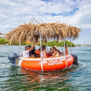 The Tiki BBQ Boat XL: Be Your Own Captain, a round orange boat with a thatched roof and outboard motor, carries six people on clear green water, surrounded by lush greenery and blue sky. Passengers chat and relax onboard.