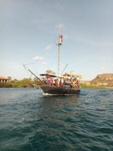 The Pirate Adventure tour boat āSimba,ā styled like a pirate ship, cruises with passengers on blue-green water past green shores, houses, and distant rocky hills beneath a clear sky. Someone stands atop the mast.