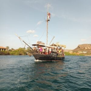 The Pirate Adventure tour boat “Simba,” styled like a pirate ship, cruises with passengers on blue-green water past green shores, houses, and distant rocky hills beneath a clear sky. Someone stands atop the mast.