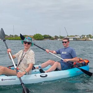 Two people in sunglasses and casual wear paddle the blue and white Tiki Kayak: Discover the Spanish Waters, with "TikiBoatRental Curacao" on its side. Boats and greenery surround them under a cloudy sky for a serene adventure.