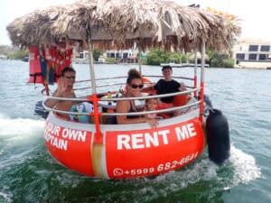 Four people, including a child, smile and wave on a round red Tiki BBQ Boat Small: Be Your Own Captain with a thatched roof and āRENT MEā sign, cruising the water with houses visible in the background.