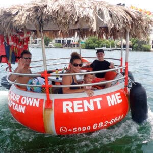 Four people, including a child, smile and wave on a round red Tiki BBQ Boat Small: Be Your Own Captain with a thatched roof and “RENT ME” sign, cruising the water with houses visible in the background.