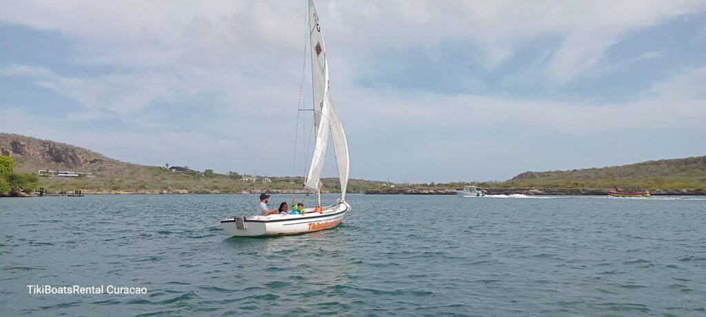 A small white Tiki Sailboat: The Ultimate Curacao Sailing Experience sails with four people on calm blue water near a green, hilly Curacao shoreline. Other boats appear in the distance under a partly cloudy sky. Text reads TikiBoatsRental Curacao.
