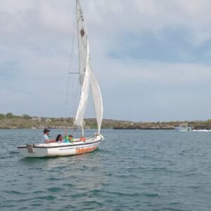 A small white Tiki Sailboat: The Ultimate Curacao Sailing Experience sails with four people on calm blue water near a green, hilly Curacao shoreline. Other boats appear in the distance under a partly cloudy sky. Text reads TikiBoatsRental Curacao.