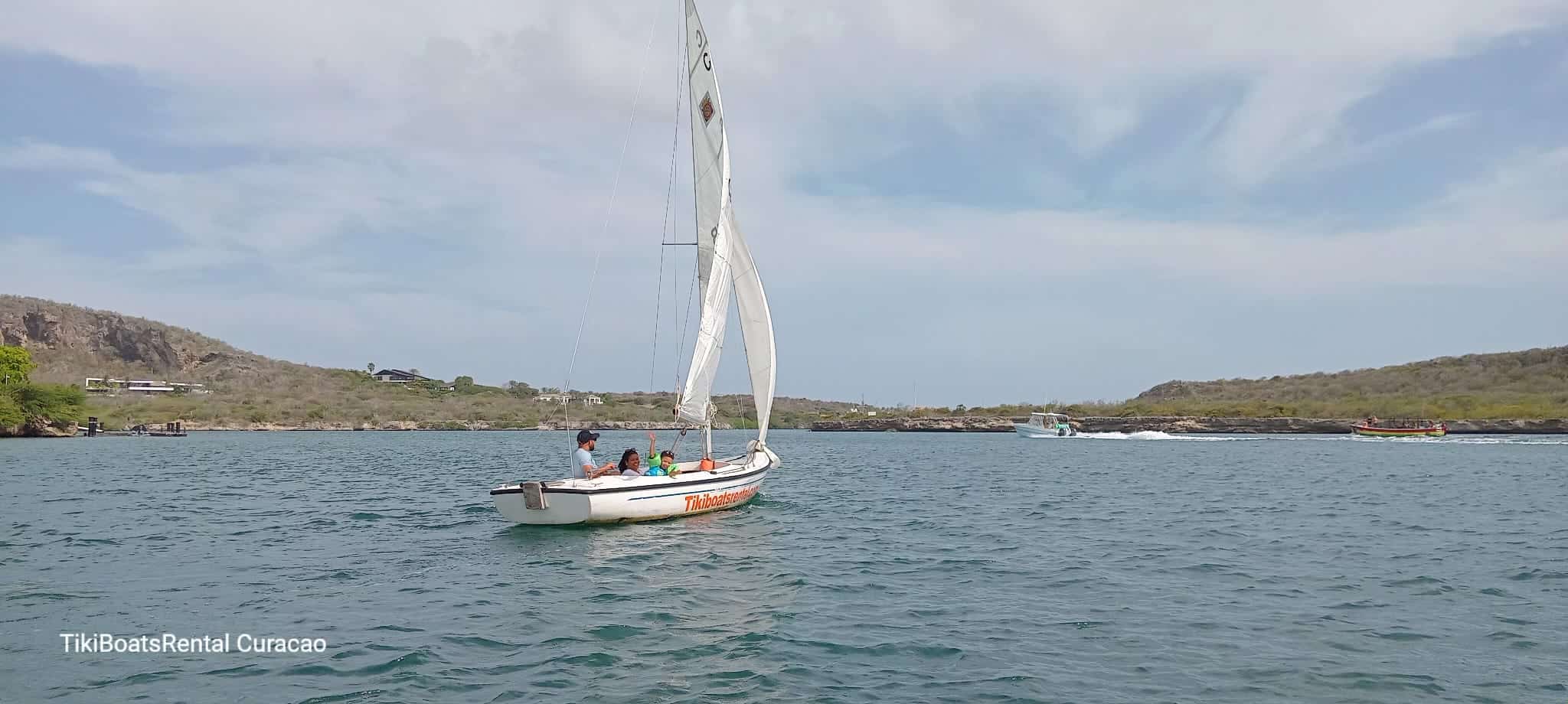 A small white Tiki Sailboat: The Ultimate Curacao Sailing Experience sails with four people on calm blue water near a green, hilly Curacao shoreline. Other boats appear in the distance under a partly cloudy sky. Text reads TikiBoatsRental Curacao.