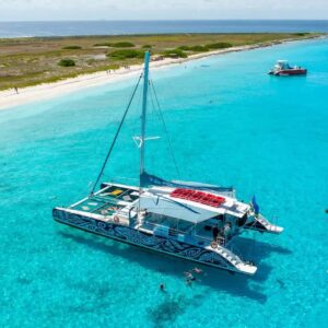 Een kleurrijke catamaran drijft op helder turkoois water nabij een zandstrand met groene vegetatie, waarbij uitgelichte producten worden getoond terwijl mensen zwemmen, snorkelen en ontspannen op boten onder een zonnige hemel. Andere boten liggen in de buurt aan de kustlijn.