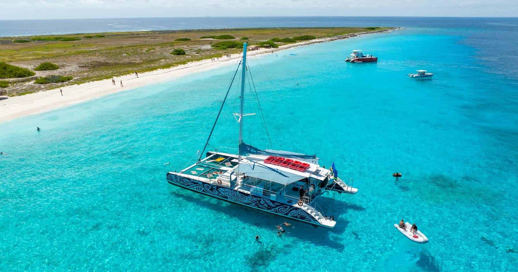 Een kleurrijke catamaran drijft op helder turkoois water nabij een zandstrand met groene vegetatie, waarbij uitgelichte producten worden getoond terwijl mensen zwemmen, snorkelen en ontspannen op boten onder een zonnige hemel. Andere boten liggen in de buurt aan de kustlijn.