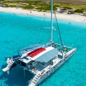 Luchtfoto van een catamaran met mensen aan boord dobberend op helder turquoise water nabij een zandstrand met zonnebaders en verspreide hutjes aan de kust.