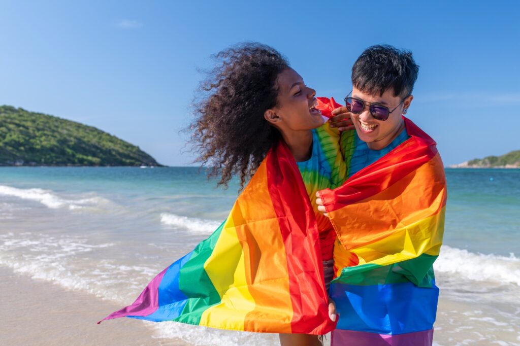 Twee mensen die glimlachen en gewikkeld zijn in een regenboog pride vlag staan op een zonnig, gay-vriendelijk strand van Curaçao met zachte golven, groene heuvels en een blauwe lucht op de achtergrond.