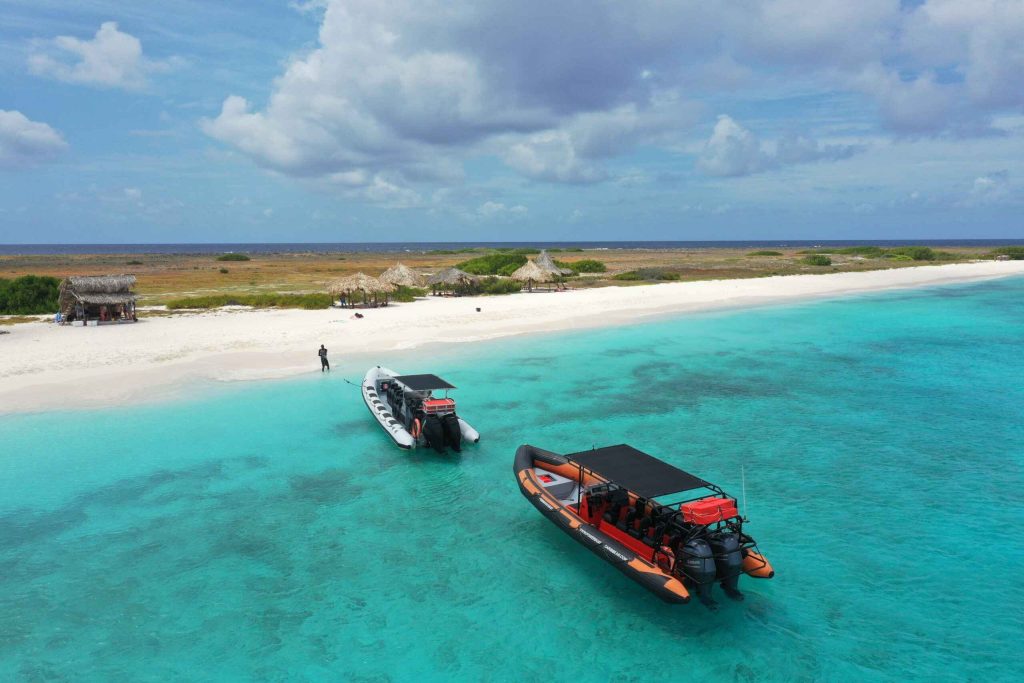 Dos lanchas rápidas flotan sobre aguas turquesas cristalinas cerca de la playa de arena de Klein Curazao, salpicada de cabañas con techo de paja y vegetación verde. Una persona está de pie en la orilla bajo un cielo azul parcialmente nublado, mientras el océano se extiende hasta el horizonte.