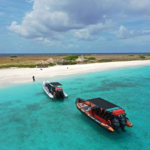 Two Powerboats float on clear turquoise water near Klein Curaçao’s sandy beach, dotted with thatched-roof huts and green brush. A person stands onshore under a blue, partly cloudy sky, as the ocean extends to the horizon.